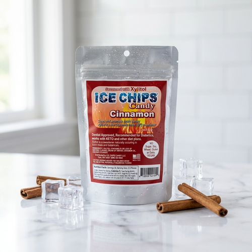 A stand-up pouch of PremiumCakes Xylitol Ice Chips Candy Cinnamon flavor, placed on a white marble surface with scattered ice cubes and cinnamon sticks. The background shows a bright, softly blurred kitchen or bathroom setting with a window.