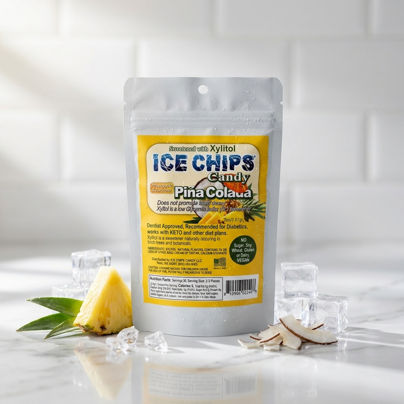 A stand-up pouch of Ice Chips Candy Pina Colada flavor, displayed on a marble surface with pineapple, coconut flakes, and ice cubes against a white subway tile background.