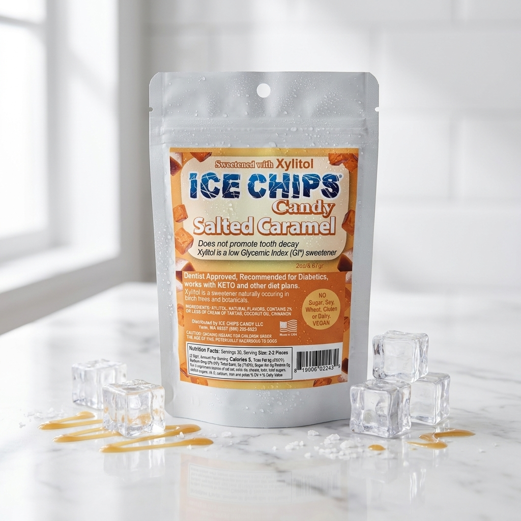 A stand-up pouch of Ice Chips Candy Salted Caramel ice chips sits on a glossy white surface with scattered clear ice cubes and drizzles of caramel around it. The package is centered against a bright, softly blurred kitchen-like background.